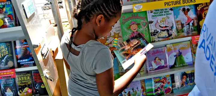 A girl in a grey T-shirt reads the back of a picture book in front of shelves of children’s books under a PICTURE BOOKS sign.