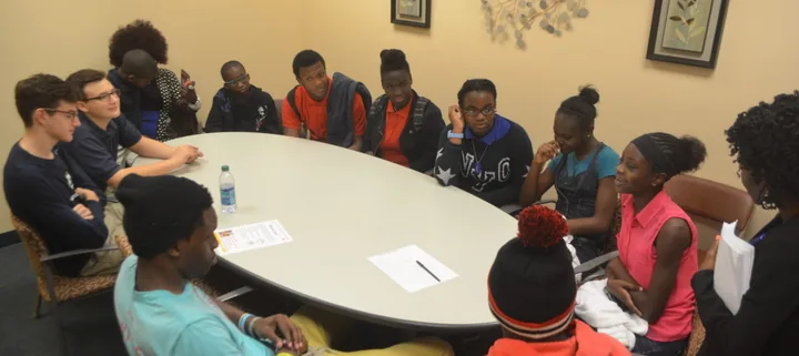 About a dozen teenagers sit around an oval conference table with papers and a water bottle; a girl in a pink top turns to speak, and a woman holding papers stands at the right edge.